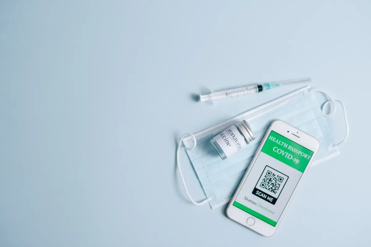 Top view of Covid-19 health passport on smartphone with vaccine, syringe, and mask on blue background.